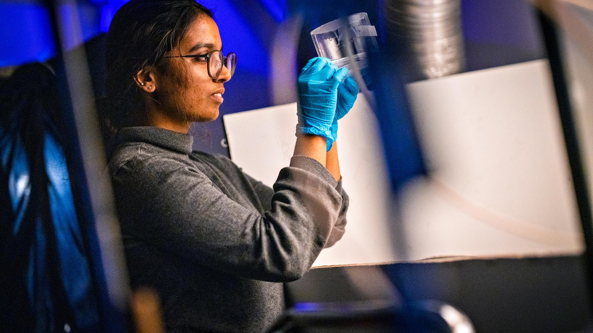 Picture of a female scientist with her side towards the camera. She wears plastic gloves and holds up a plastic container and observes its' contents.