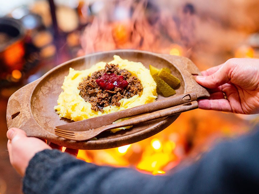 Händer håller en tallrik med renskav och potatismos invid en öppen eld. Foto.