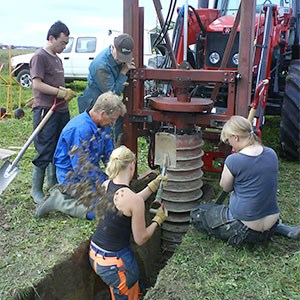 Five people at a tractor on a field, one of the people standing in a hole in the ground, photo.