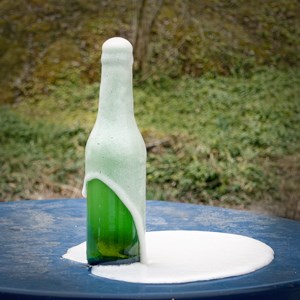 En grön flaska som har skummat över. Flaskan står utomhus på ett blått bord. Foto.