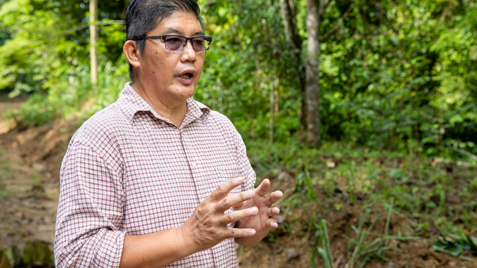 Man in shirt in rainforest talks and gestures with his hands.