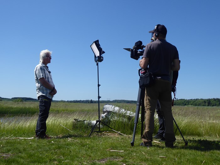 En man blir filmad av två andra, framför en våtmark. Foto.