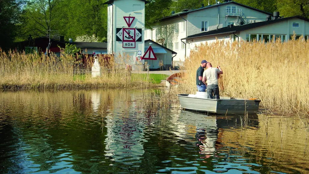 Två personer står i en liten aluminiumroddbåt vid vassen nära strandkanten. I bakgrunden syns en byggnad med vit fasad och skyltar som varnar för rör i vattnet. Vattnet speglar både båten och skyltarna, och omgivningen består av grönskande träd och vass.