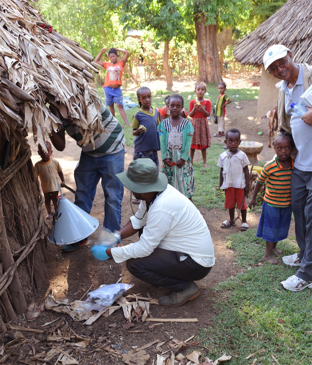 Bild av en man som monterar en myggfälla vid ett hus i byn Magge. Runt omkring har folk samlats för att titta på.