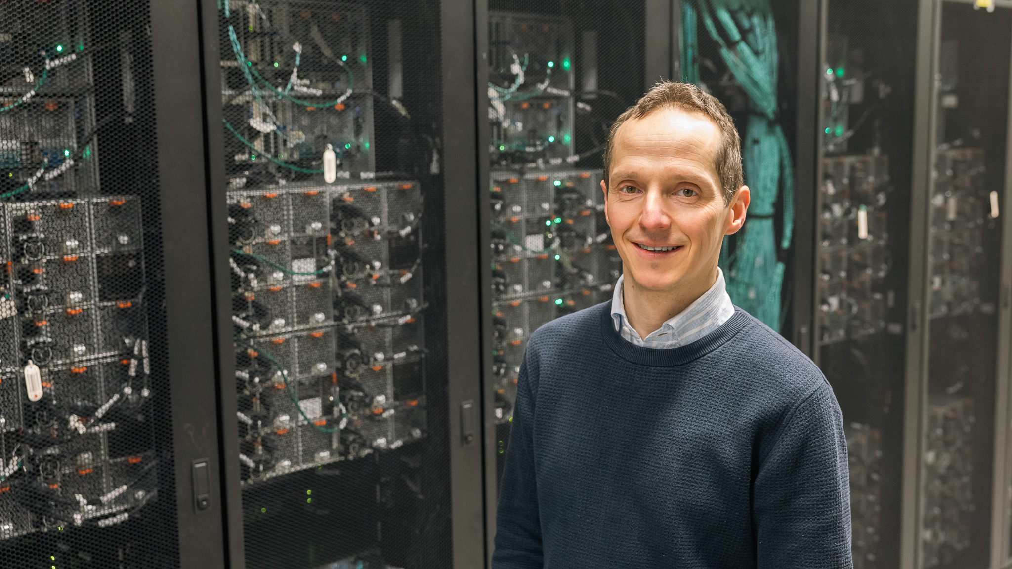 Portrait of Nicolas Delhomme, coordinator of the WIFORCE Data Support Centre, standing in front of server racks in a data centre. Servers, network cables, and green status lights are visible in the background, illustrating the technical infrastructure used for data management and computing.