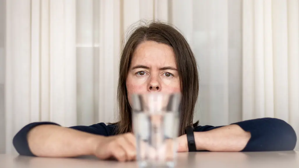 Portrait of a woman (Carolina Vogs, SLU) looking straight into the camera. In the foreground is a water glass through which the woman is looking.