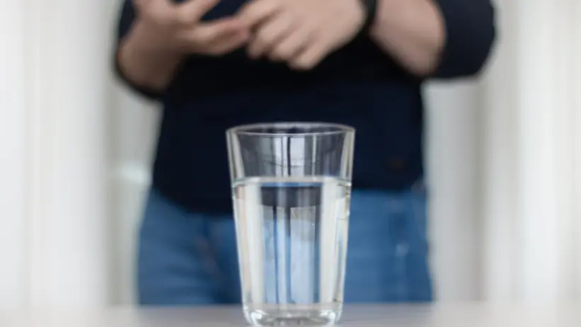 Photo of a water glass on a table, with a woman gesturing with her hands (Carolina Vogs, SLU) in the background.