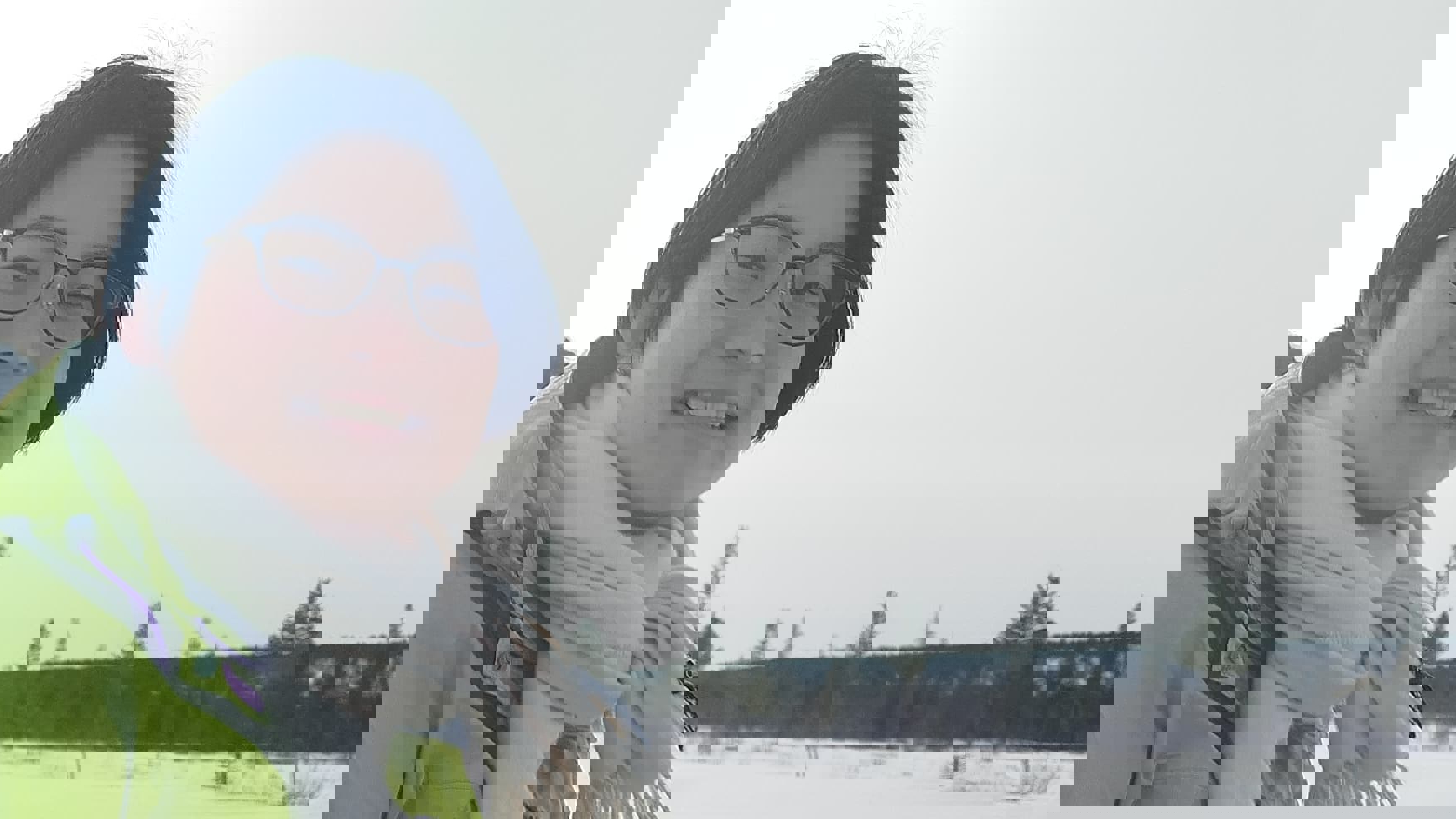 A smiling person in a green jacket and white scarf sitting outdoors in winter, with snow and other people in the background.