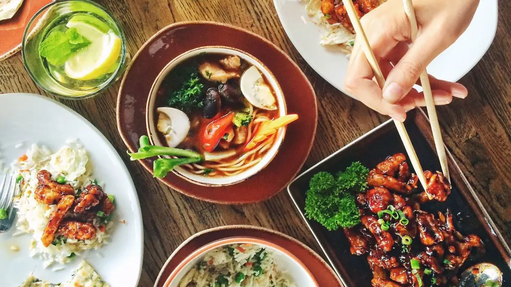 Fat med asiatiska maträtter och en hand med ätpinnar. Foto.