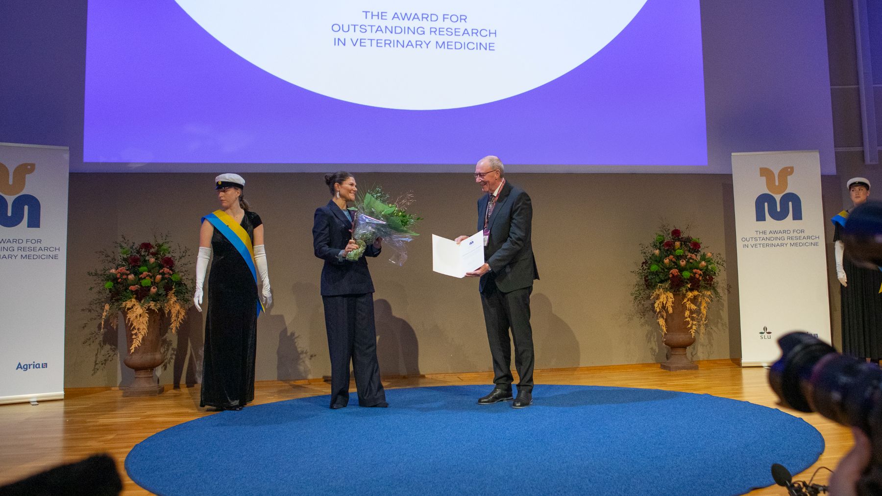 A formal award ceremony on stage, where a woman in a suit hands a bouquet to a man receiving an open certificate. Two ceremonial attendants stand on either side.