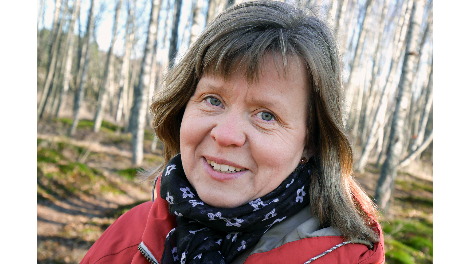 Mörkblond kvinna i röd jacka och svart scarf med vita blommor står i björkskog. Foto