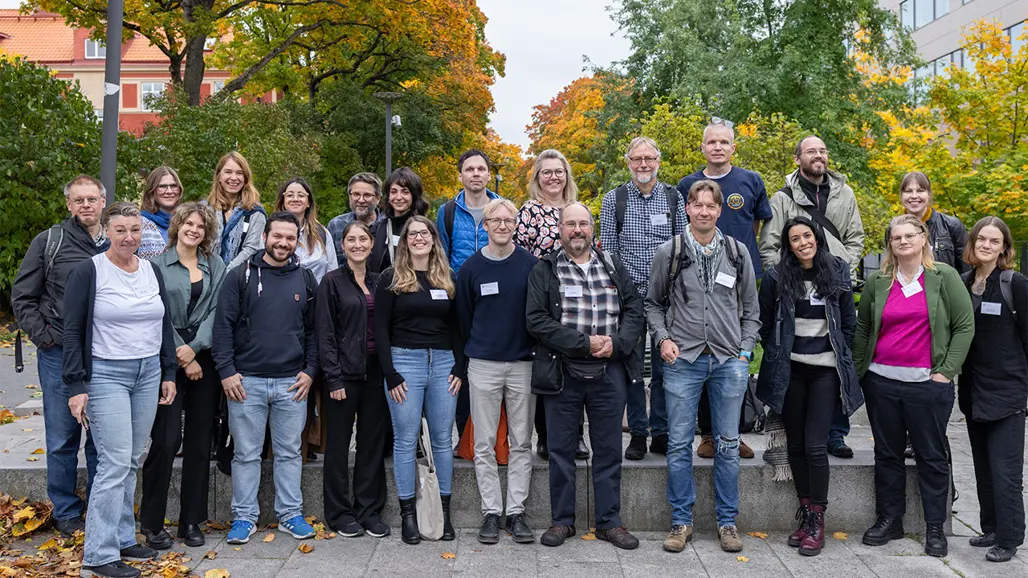 23 personer står uppradade för gruppbild utomhus framför höstfärgade lövträd omgärdade av flervåningshus. Foto