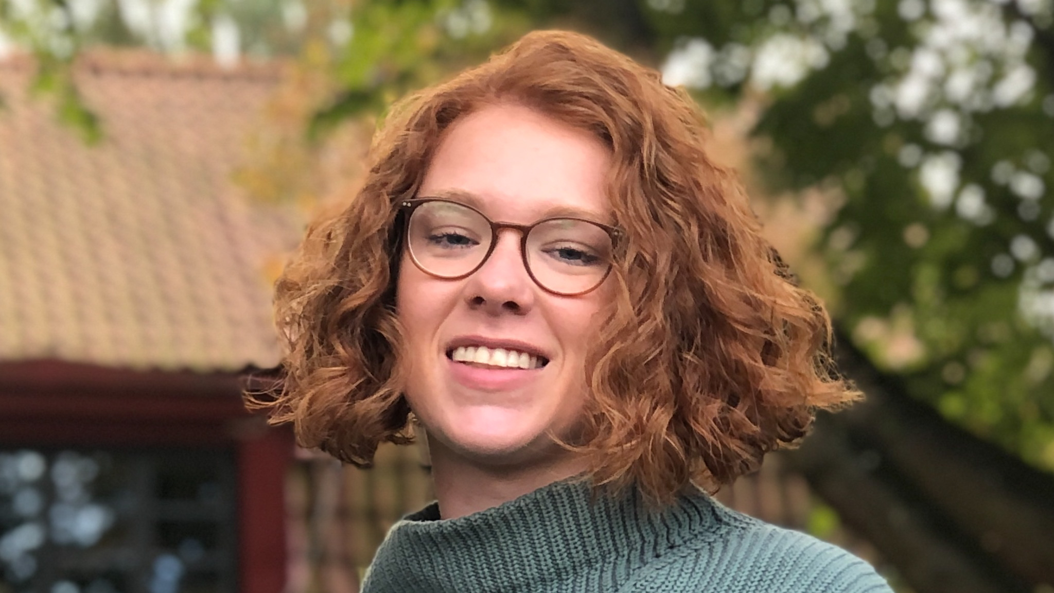 Close-up of a person with shoulder-length curly hair and glasses. The person is wearing a ribbed knit jumper and is outdoors with a tiled roof and green trees in the background.