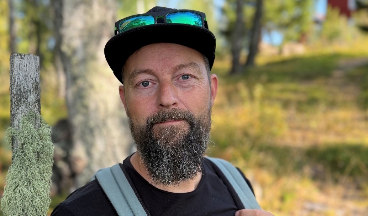 Porträttfoto av en person utomhus i skogsmiljö. Personen bär svart keps med solglasögon ovanpå skärmen, svart tröja och en ljusgrå ryggsäck. Personen har skägg och tittar mot kameran.