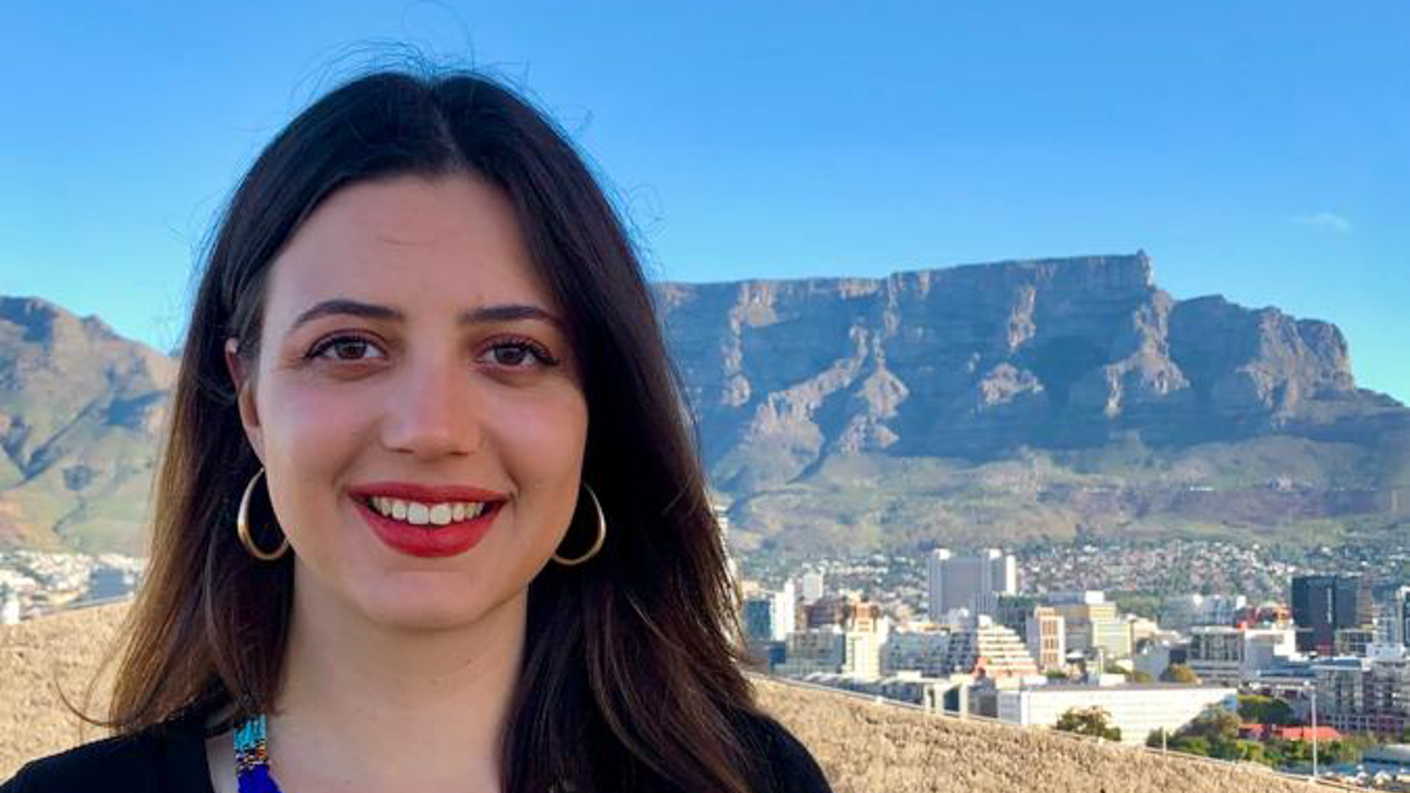 A smiling person with long dark hair, a colourful beaded necklace, and large earrings stands in front of a city with a distinctive flat-topped mountain in the background, on a clear day.