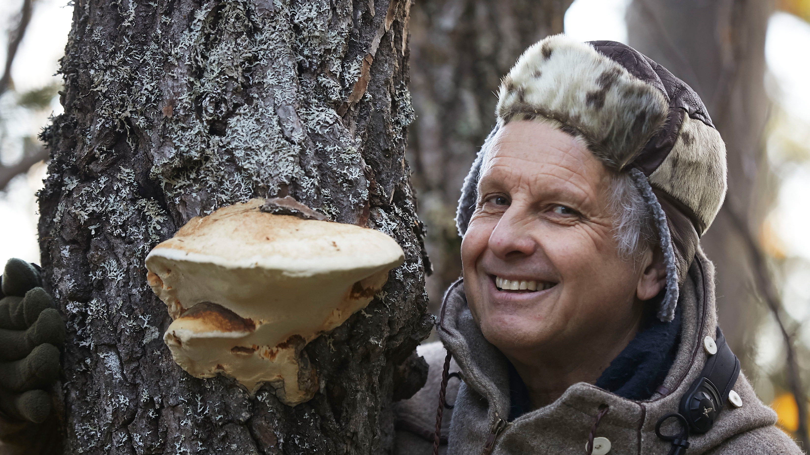 Glad man med grått hår och vintermössa står vid ticka på trädstam. Foto