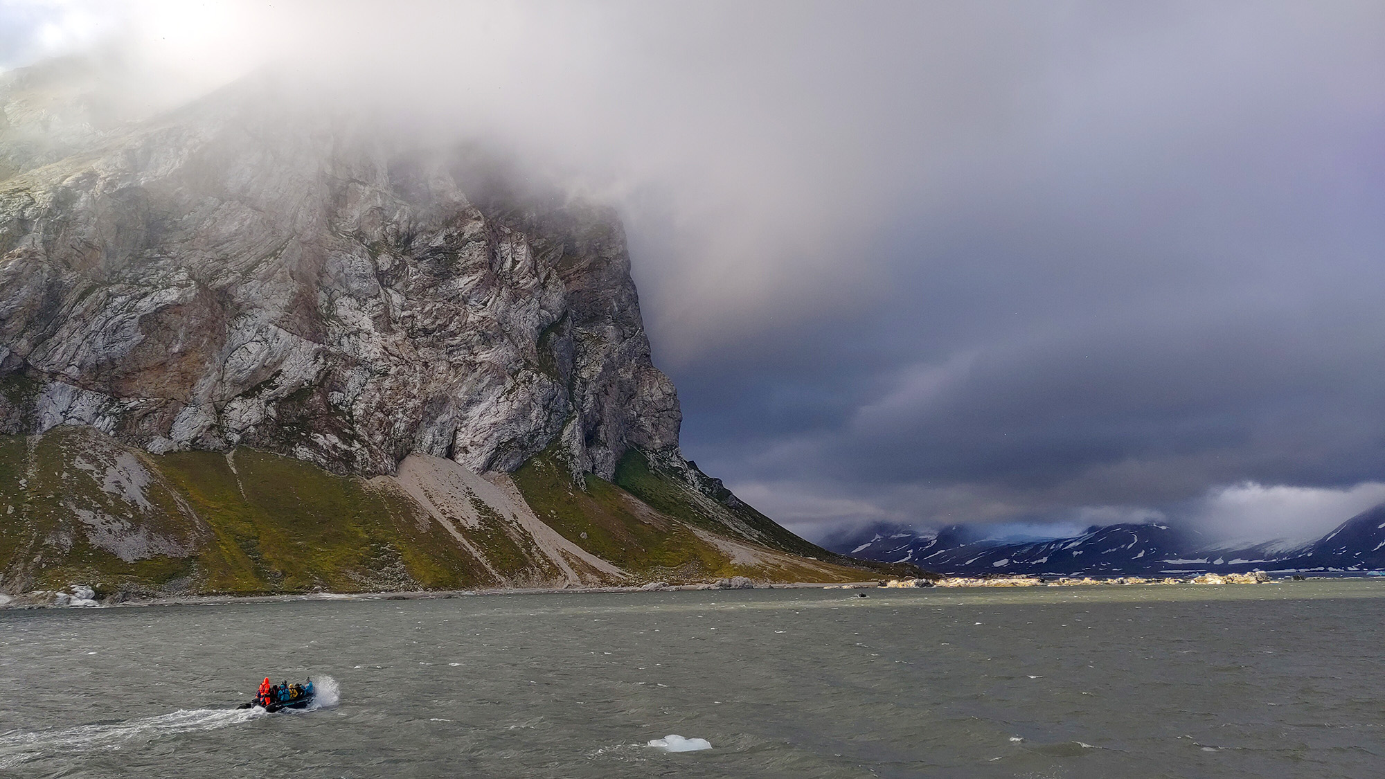 Liten gummibåt med personer i röda flytdräkter kör på ett grått hav nedanför ett brant, dimhöljt berg i ett arktiskt landskap.
