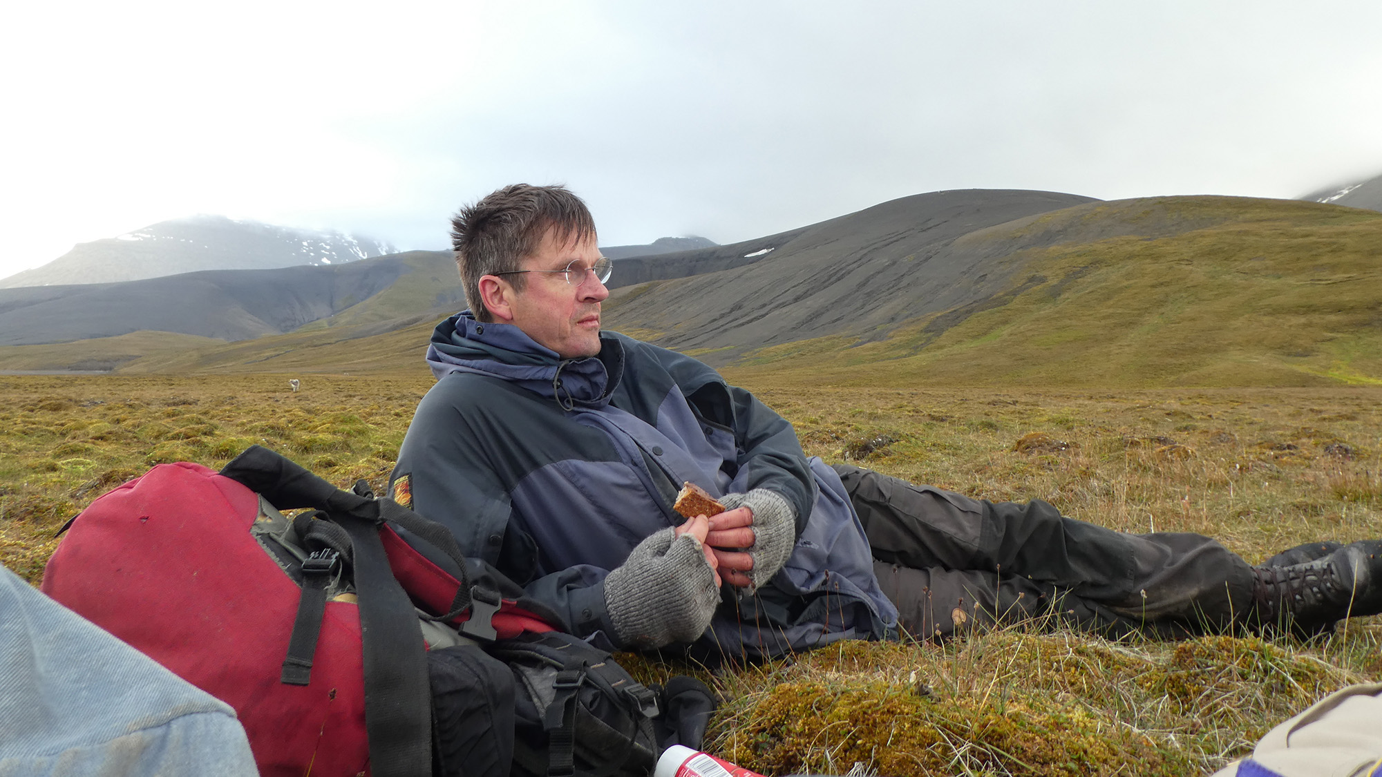 Man i friluftskläder ligger på marken i ett öppet, kargt landskap med ryggsäck bredvid sig och håller i en smörgås.