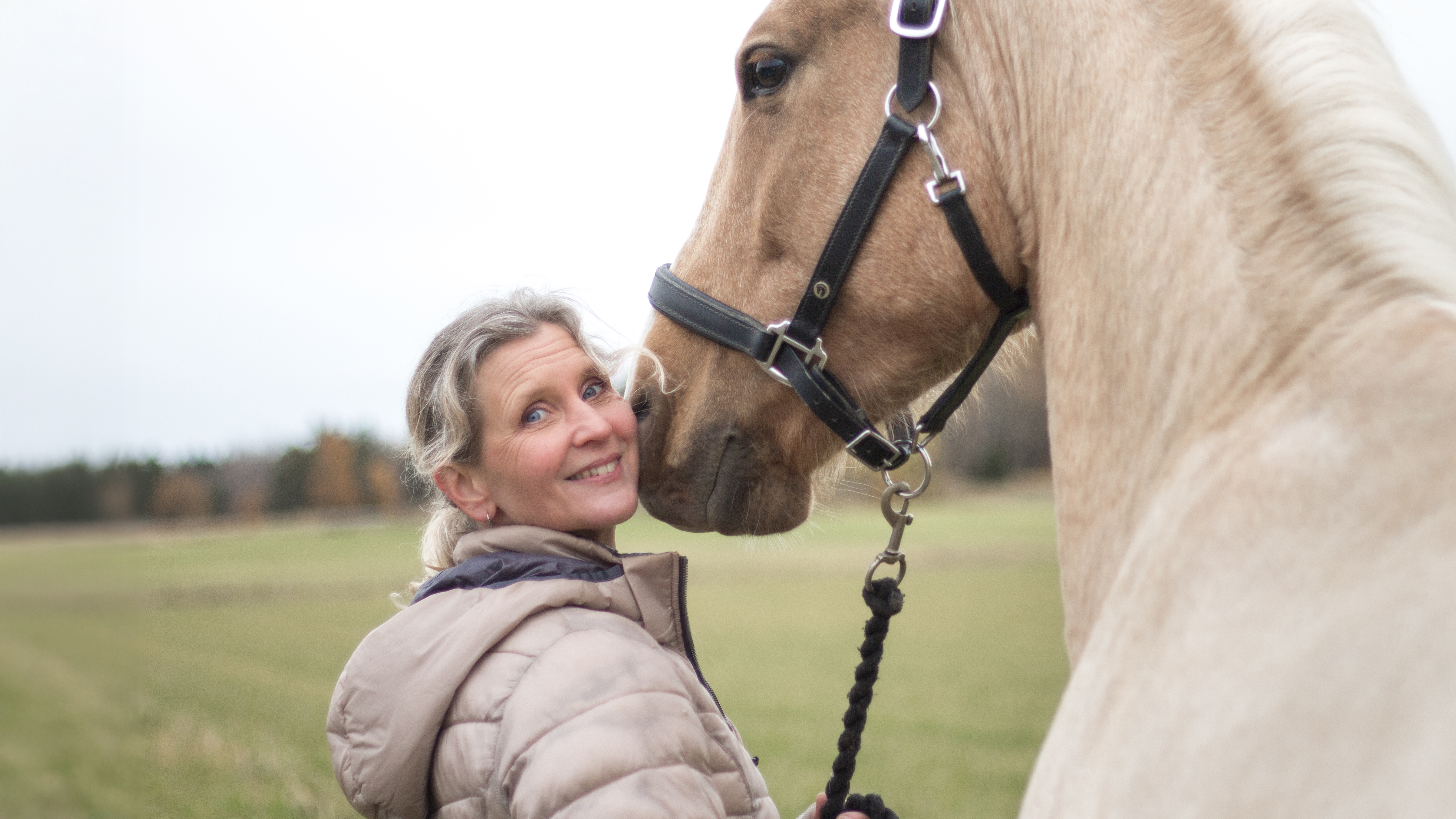 Porträttbild av en kvinna (Åsa Gelinder Viklund) ute på en äng tillsammans med en häst.