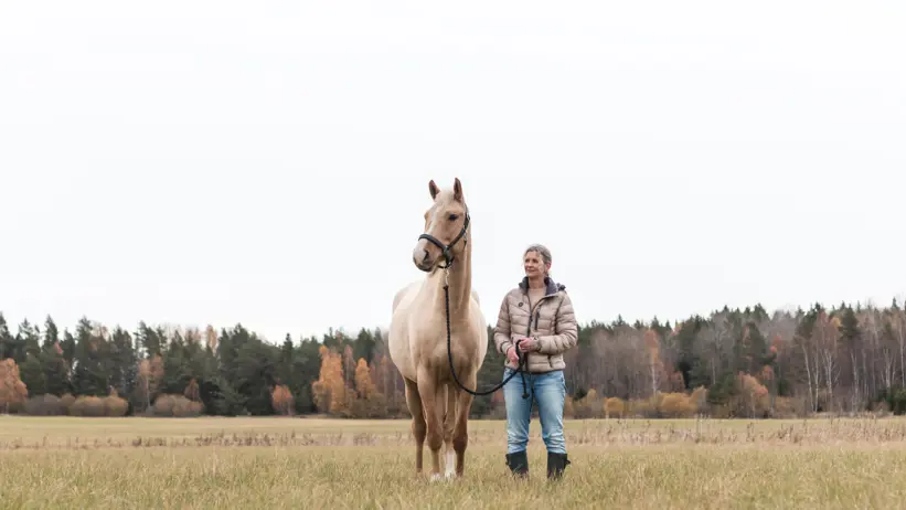 Foto av en kvinna (Åsa Gelinder Viklund) ute på en äng tillsammans med en häst.