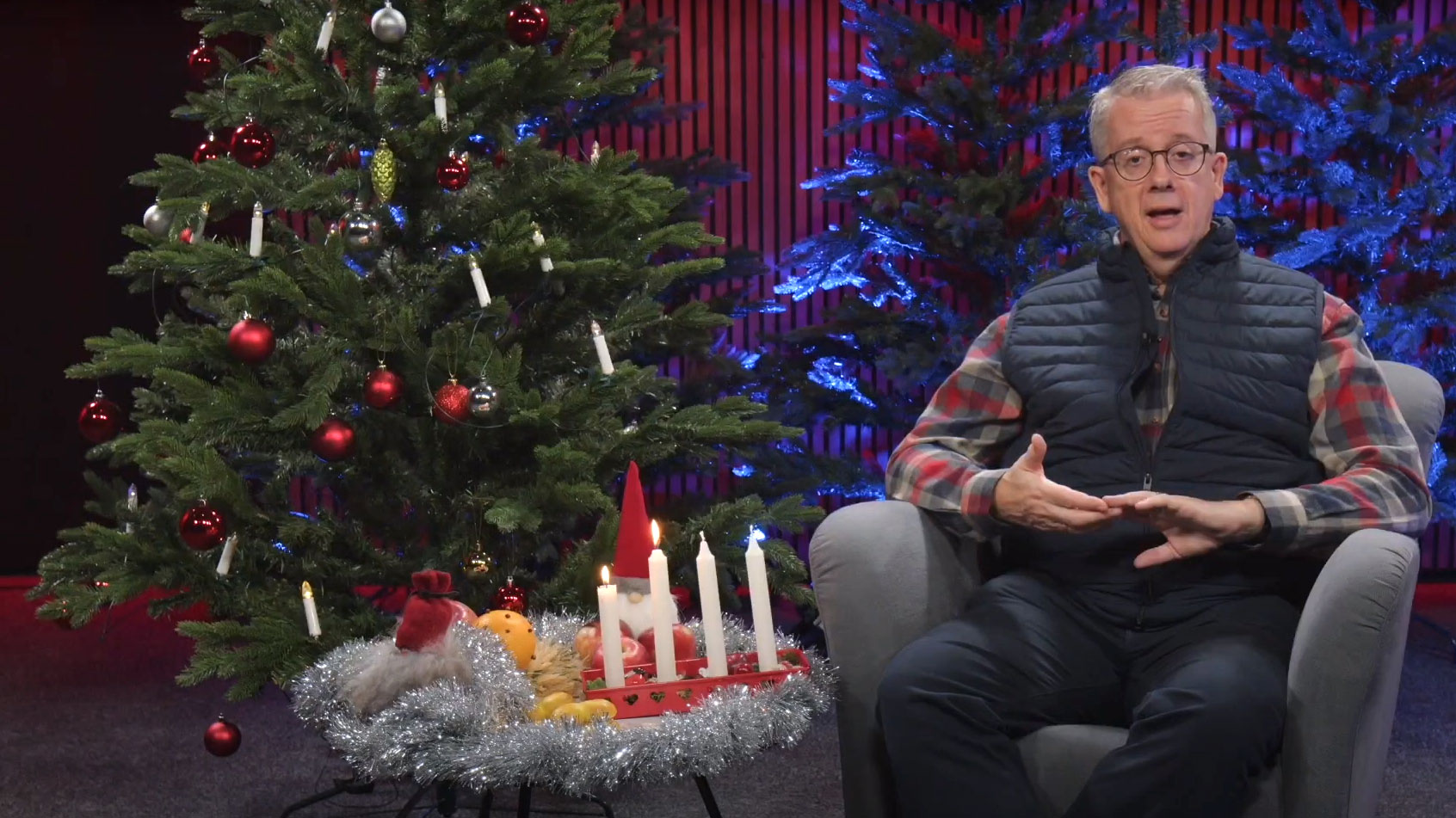 En man sitter i en fåtölj bredvid en julgran. På bordet en adventsljusstake och lussebullar. Foto.