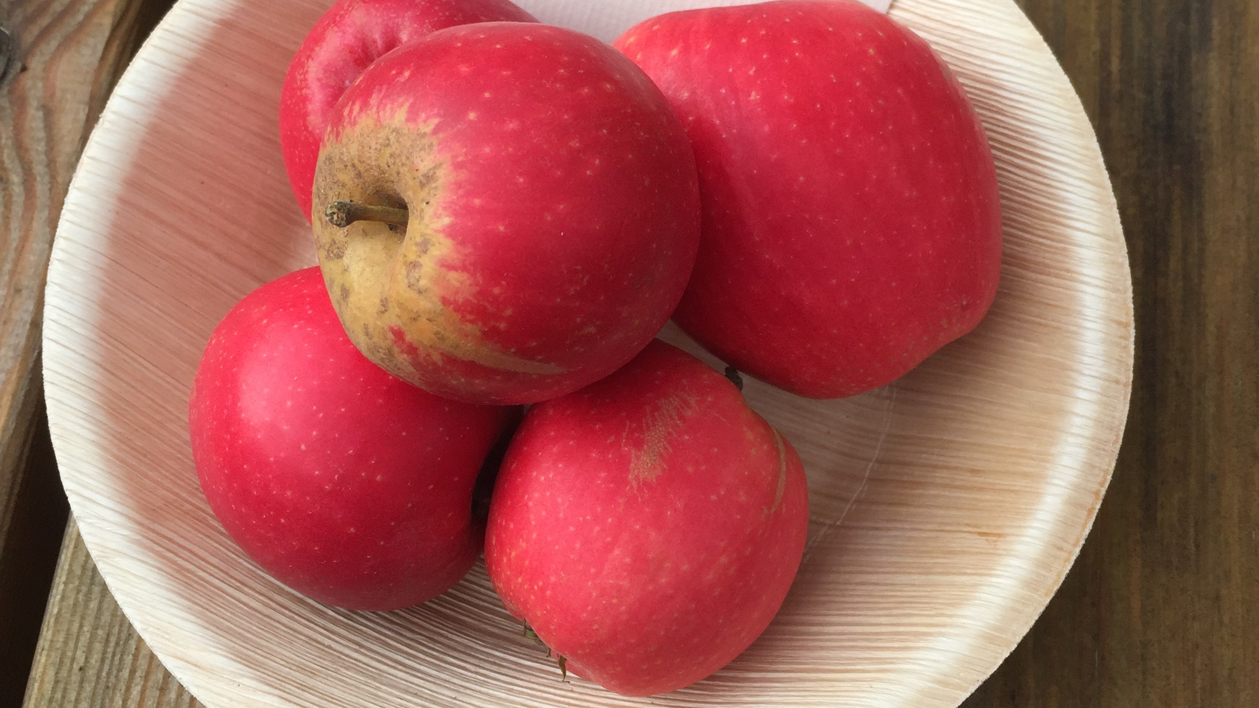 Närbild av fyra röda äpplen som ligger i en träskål. Skålen står på ett bord av trä. 