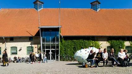 Studenter utanför en tegelbyggnad med rött tegeltak och grönskande växter på fasaden på Campus Alnarp, sittande och promenerande på en solig innergård med en vit skulptur med röda prickar i förgrunden.