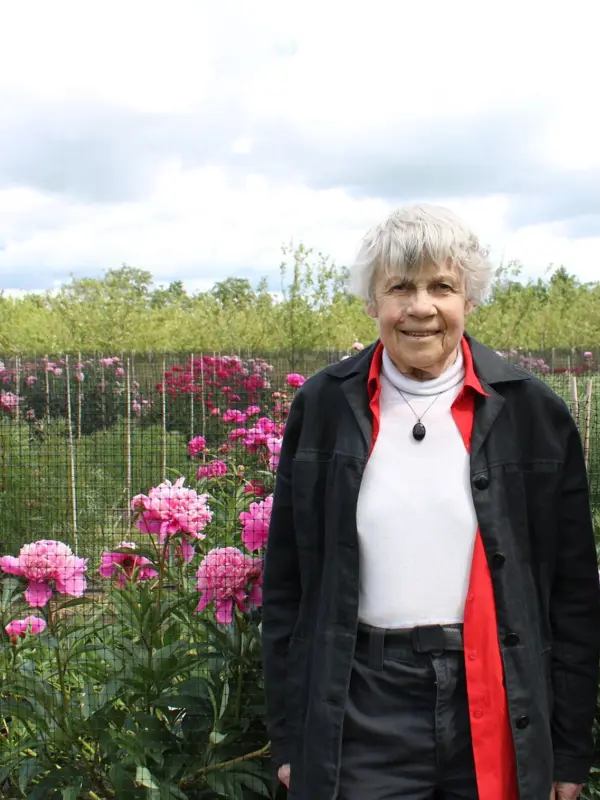 Essie Kjällquist står i genbankens perennsamling. Hon har en mörkt blå ytterjacka och står framför rosa luktpioner.