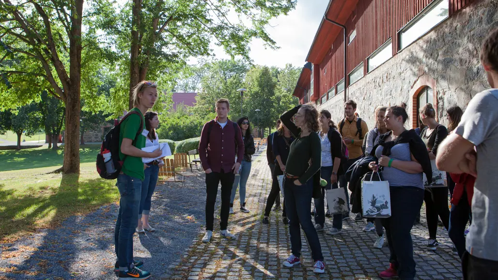 En grupp människor står utomhus på en kullerstensgång nära en röd byggnad med stenväggar. Några håller i papper eller kassar, och bakgrunden visar grönska med träd och gräs under en klar himmel.
