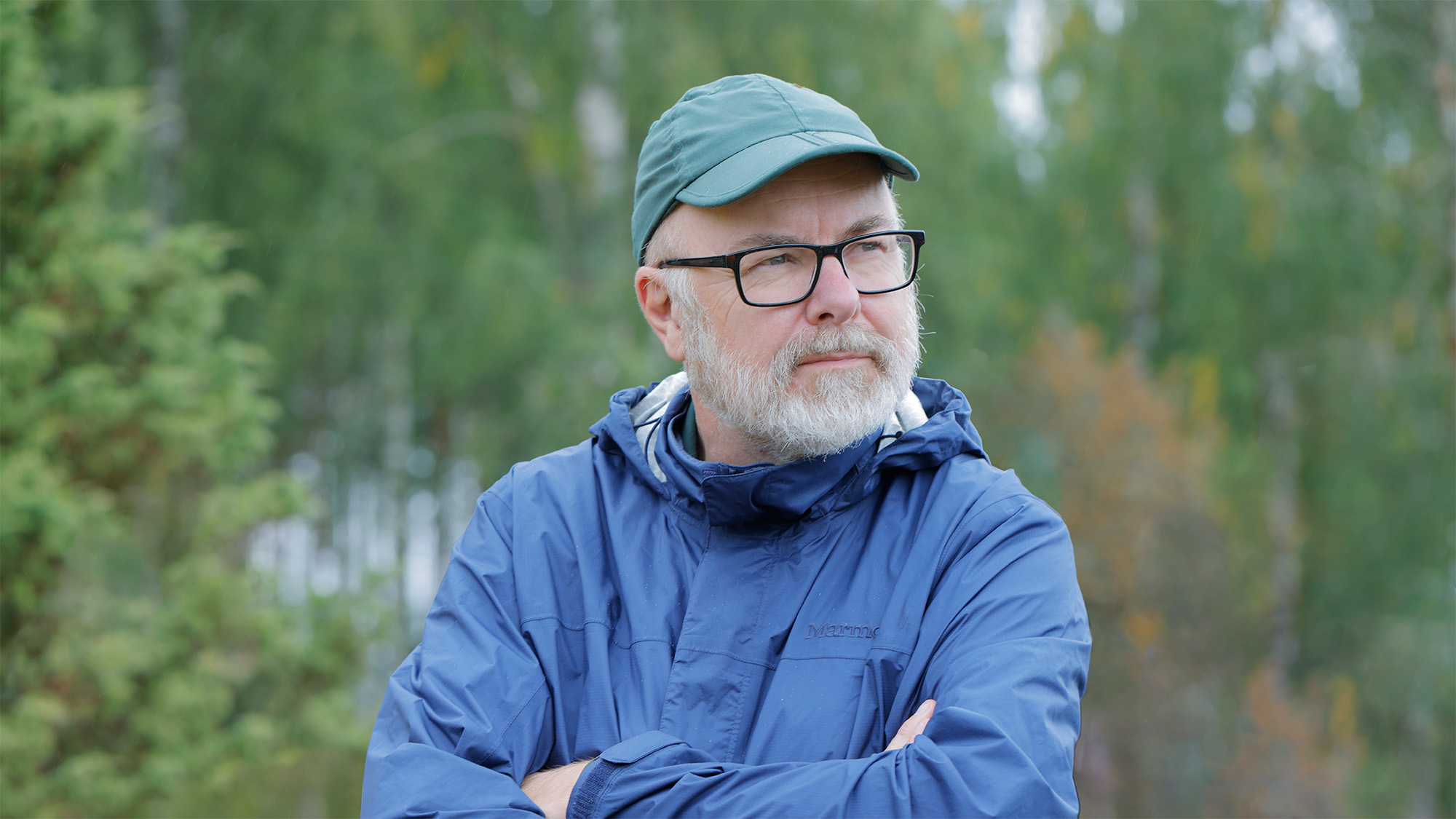 Man med glasögon, vitt skägg, blå regnjacka och grön keps står med armarna i kors i skogsmiljö. Foto