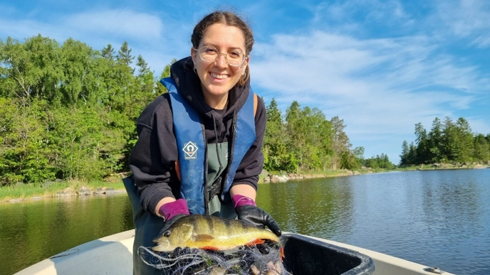 A smiling person wearing goggles and a life jacket holds a large perch in a net on board a small boat. In the background, a calm lake surrounded by lush trees under a clear blue sky.