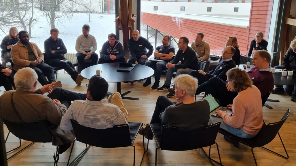 Nineteen people are sitting on chairs in a circle around a table.