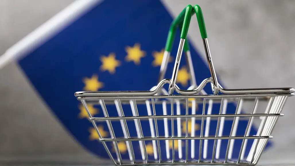 En kundkorg i miniatyr framför en EU-flagga. Foto: Shutterstock.