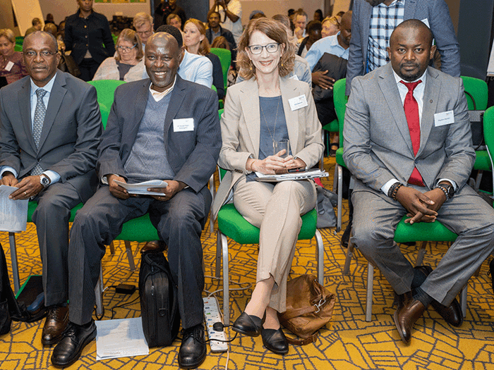 A woman and three men, all of them wearing suits and with name tags on their chests, are sitting on green chairs in a row, looking into the camera. 