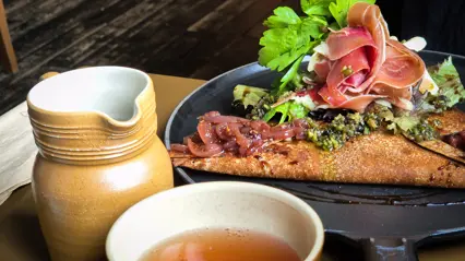 En ciderkanna och cidermugg i lera bredvid en galette med skinka