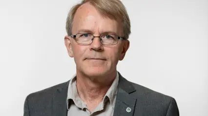 En person med ljust, kort hår och glasögon står mot en neutral bakgrund. Personen bär en grå kostym med en ljus skjorta och en liten pin på kavajen.