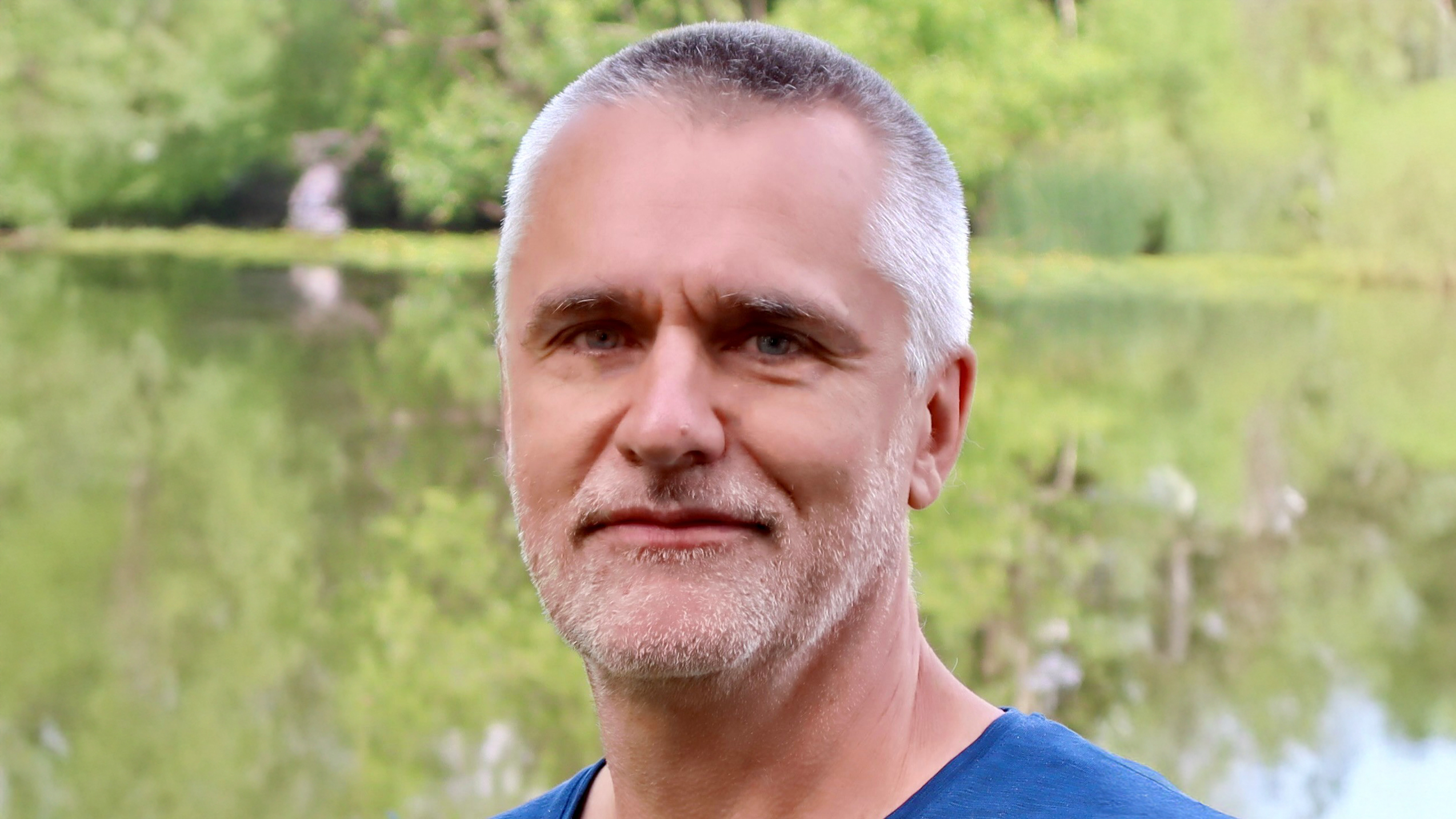 Portrait of a man with short grey hair and a beard, outdoors in front of a lake and greenery.