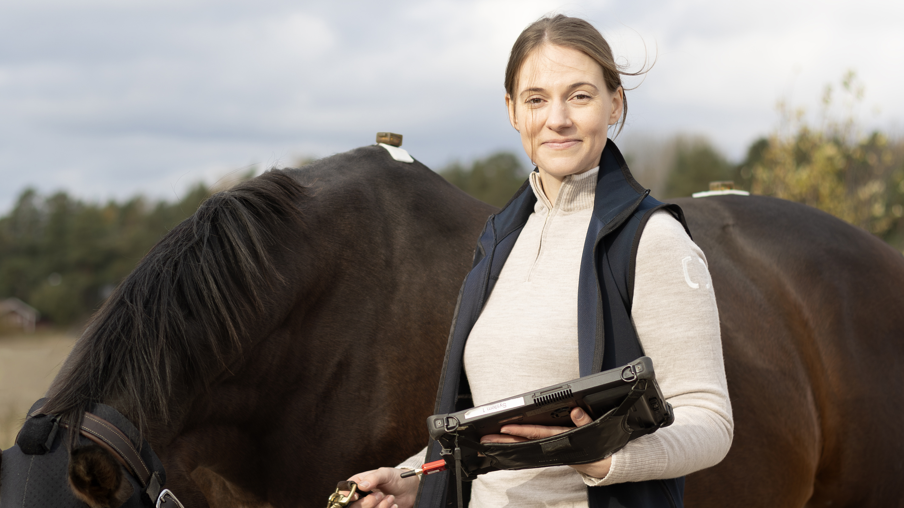 Foto av en kvinna (Ebba Zetterberg) som står utomhus bredvid en stor brun häst med en elektronisk platta i handen.