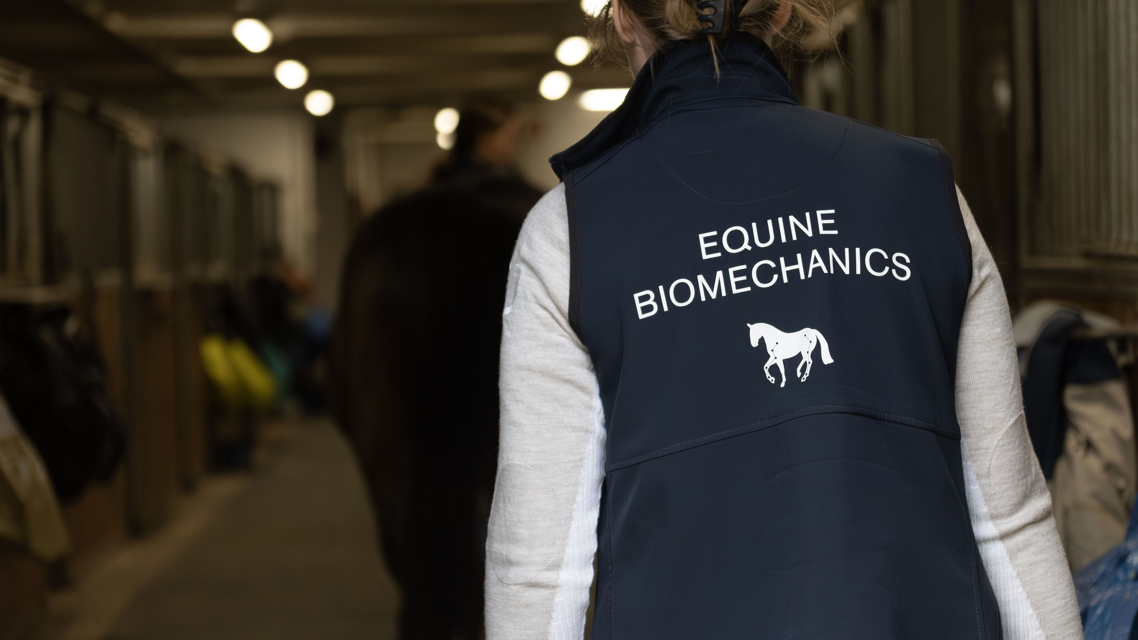 Foto av en kvinna i väst med texten "Equine biomechanics" i stallmiljö.