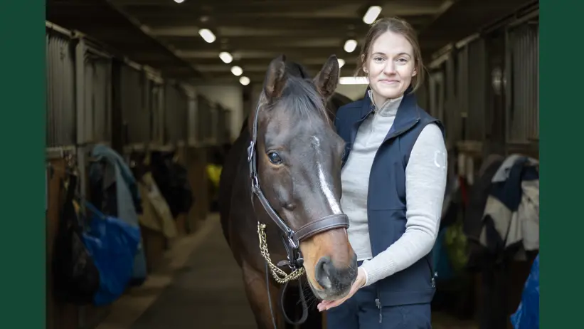 Portrait photo of a woman (Ebba Zetterberg, SLU) with a big dark brown horse in a stable setting.