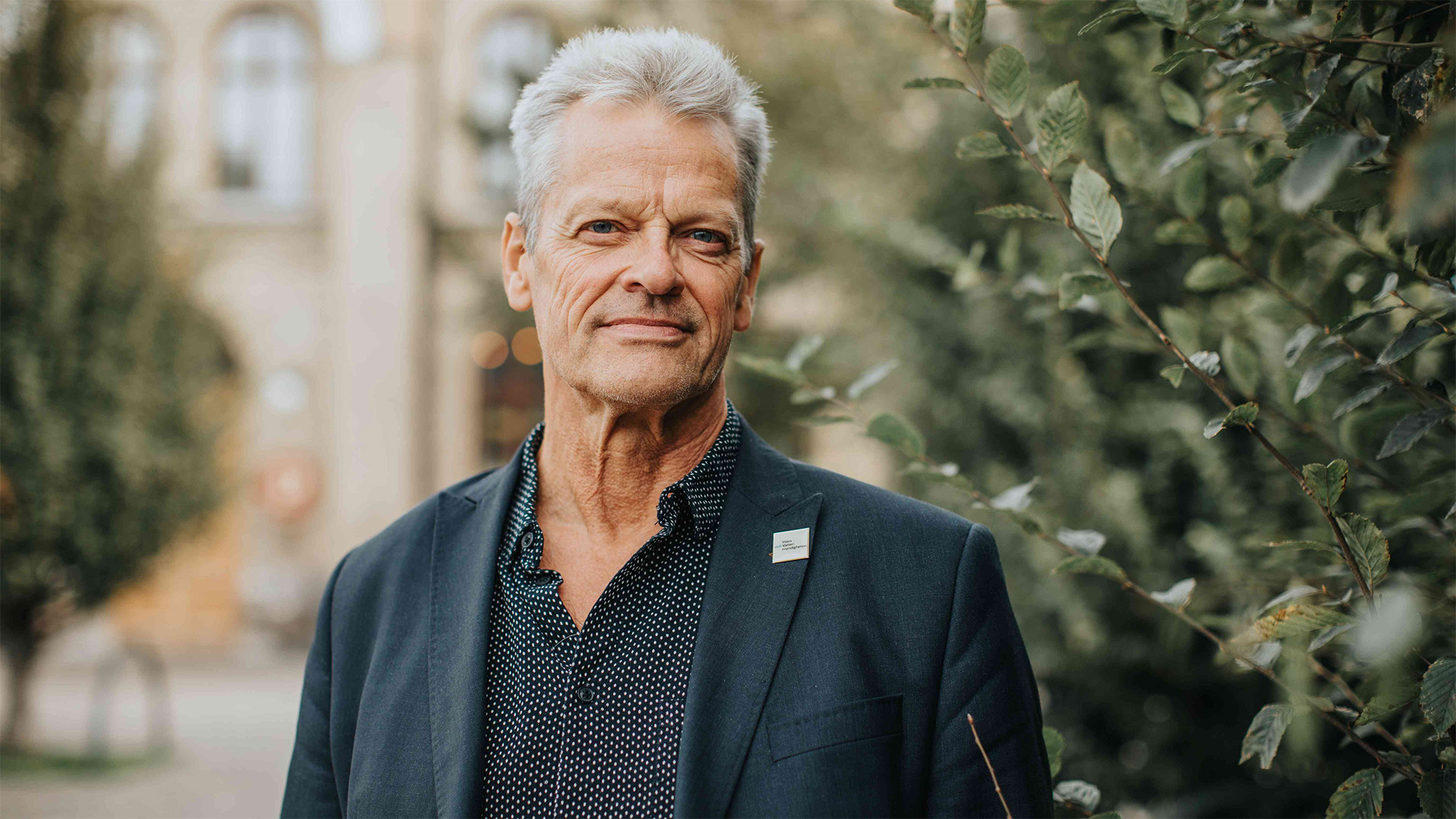 Slätrakad man med grått hår och marinblå kavaj står framför gammal byggnad. Foto