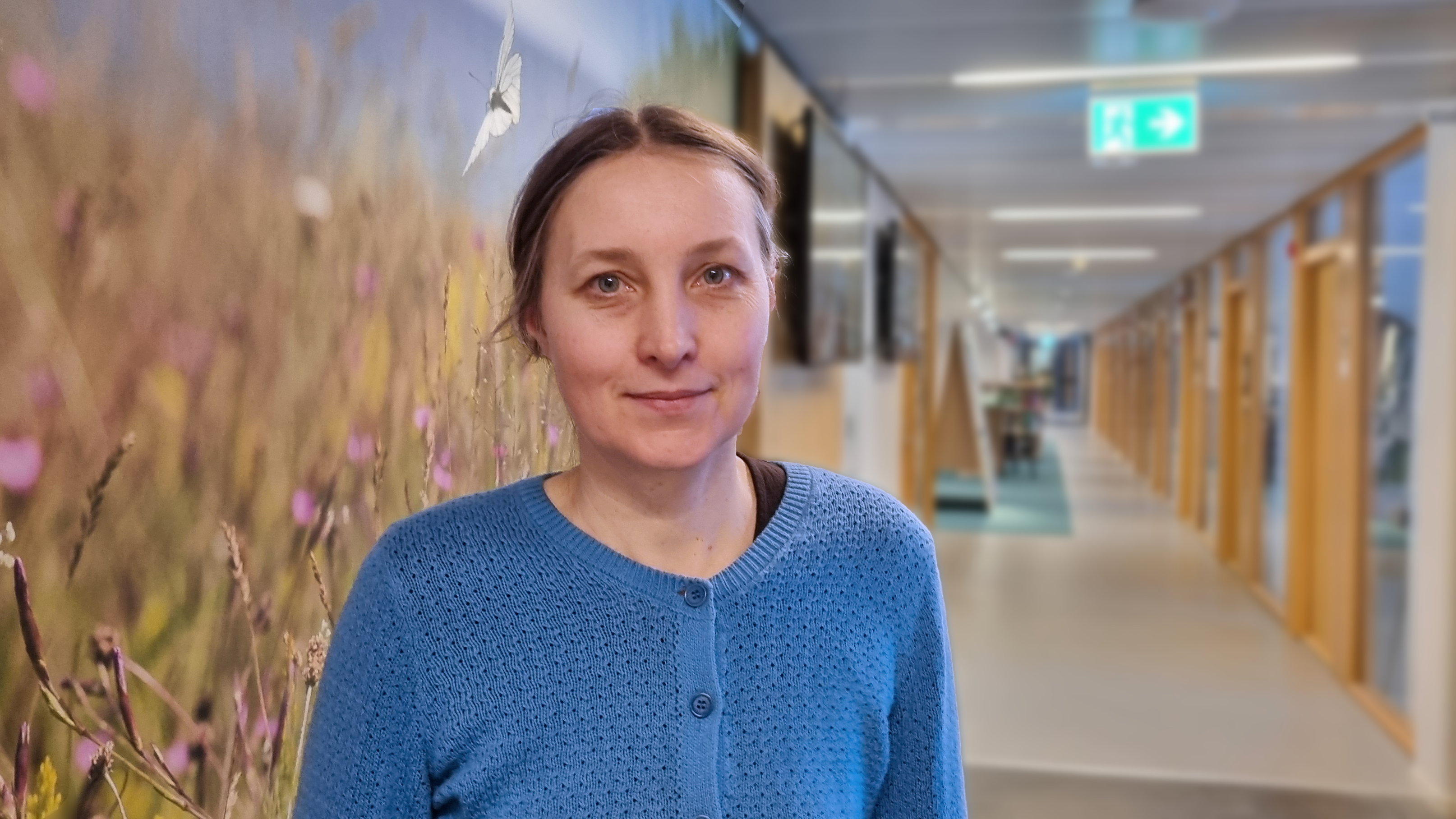 Leende kvinna med brunt, uppsatt hår och blå kofta står i korridor. Foto