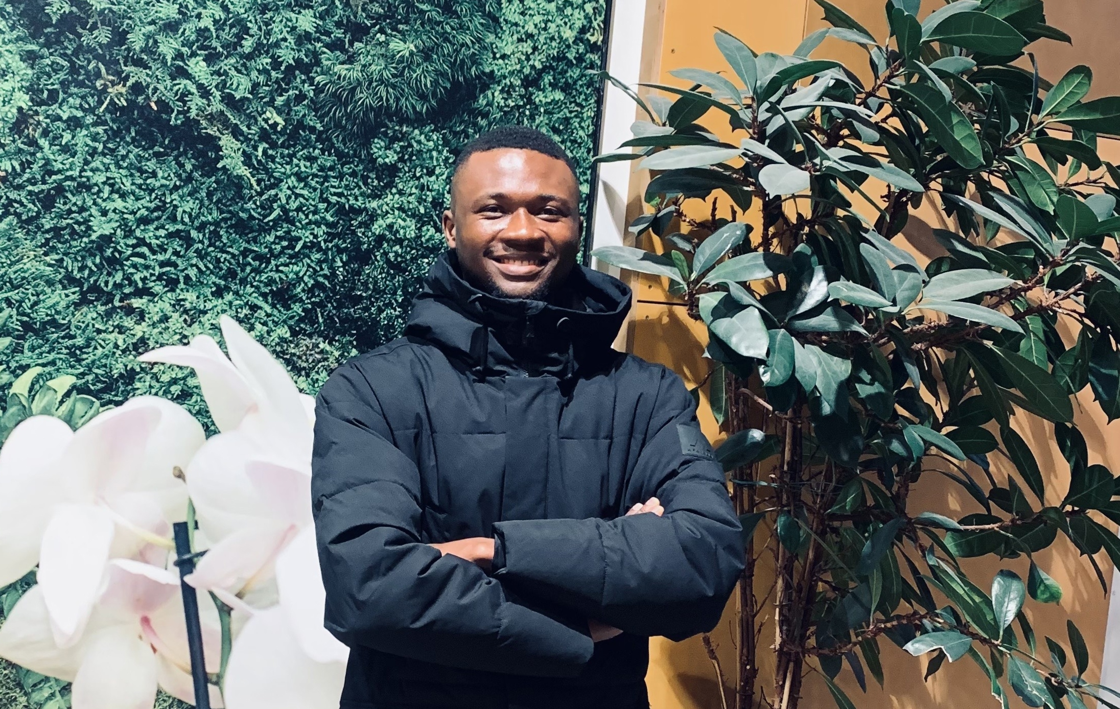 Wisdom, an SLU student ambassador, smiles at the camera in front of a green plant wall.