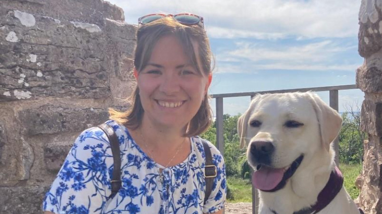 Porträttbild på Kajsa Lätt med en hund bredvid sig. Stenmur och blå himmel i bakgrunden.