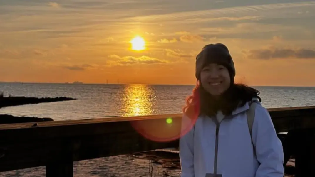 A person wearing a white jacket and beanie smiles at the camera by a pier, with the sea and a warm sunset behind them. The sun reflects on the water.