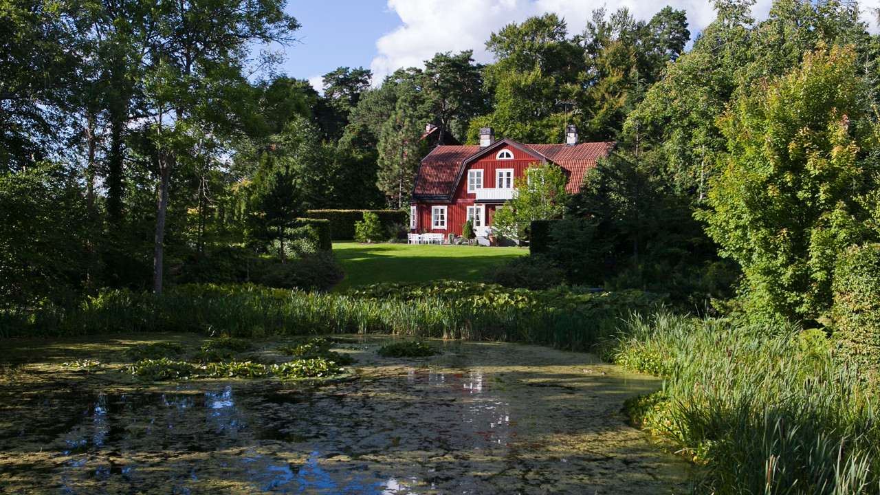 A red building surrounded by greenery with a pond in the foreground. Photo.