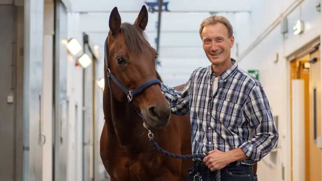 Photo of a man in a checked shirt (Tomas Bergström, SLU) standing next to a brown horse in a stable.