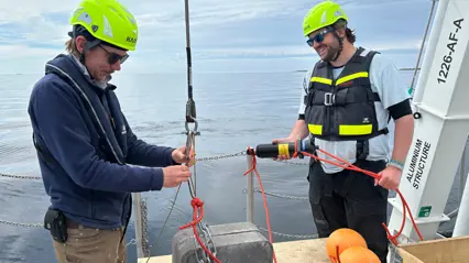 Two people in safety gear work with ropes and equipment on a boat deck over calm water, preparing scientific instruments for deployment.
