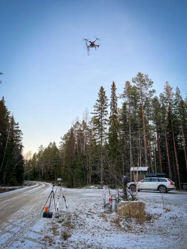 Drone flying above a forest road with measurement equipment set up on the ground next to a parked car.