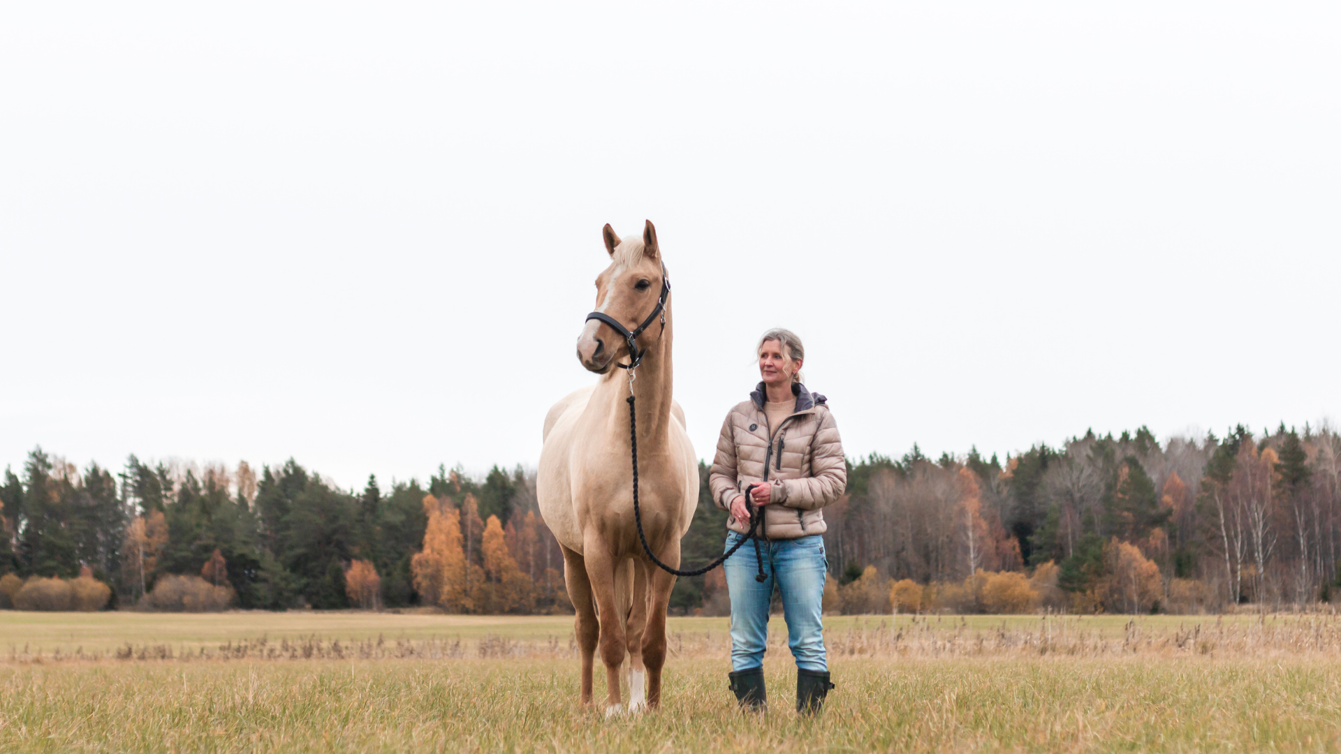 A woman wearing a beige jacket and blue jeans stands in a field with a light brown horse next to her. Behind the woman and the horse are green and orange trees.